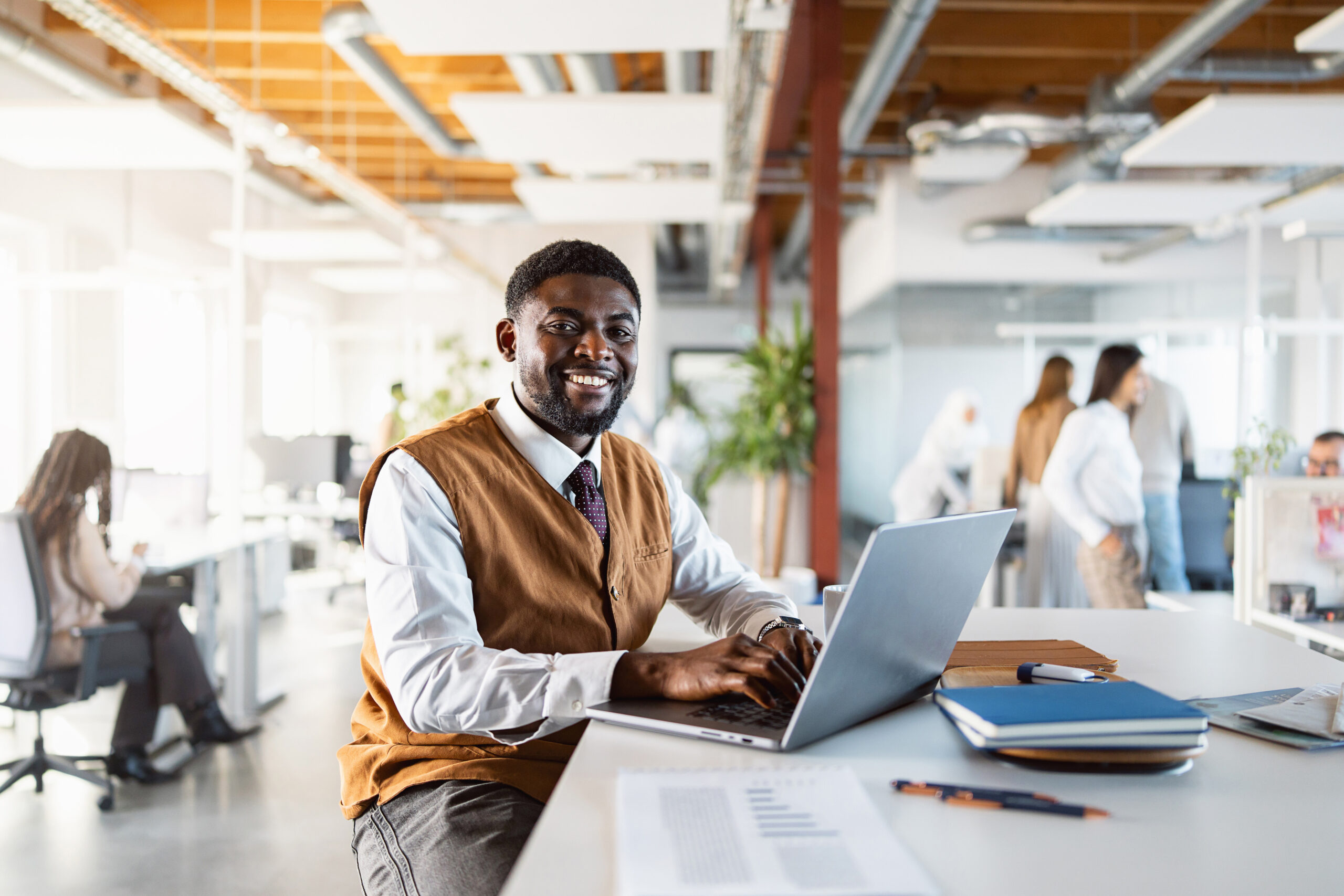 A confident businessman is seated at a desk in a modern office, working on a laptop. He is dressed in a smart casual outfit and appears engaged in his work, with colleagues visible in the background discussing ideas and collaborating.